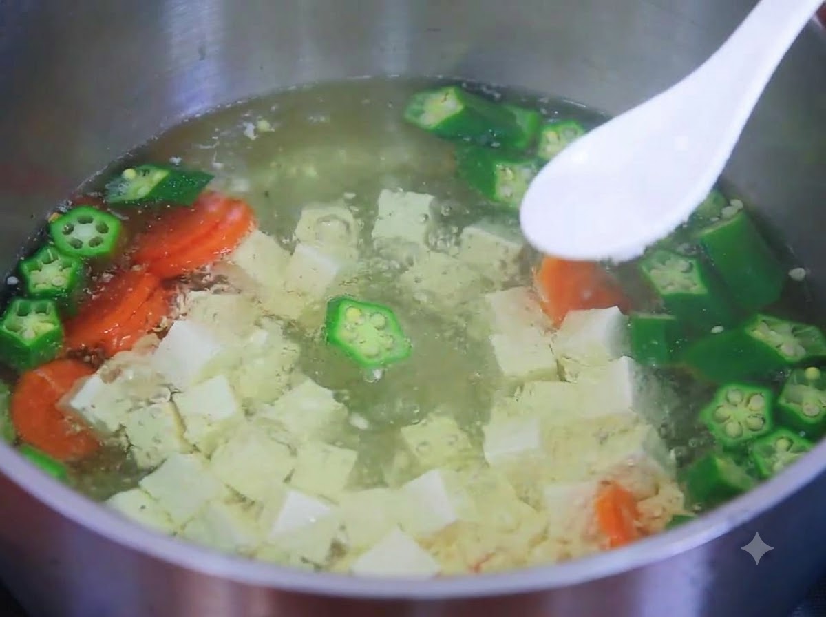 A white spoon holding salt over a simmering pot of tofu, okra, and carrot soup.