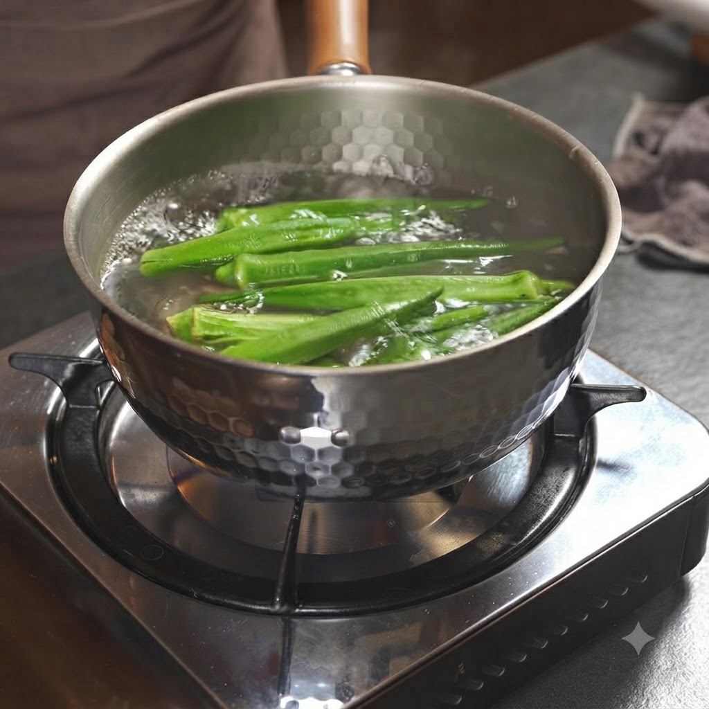 Green okra pods boiling inside a metallic pot filled with water on a stovetop burner.