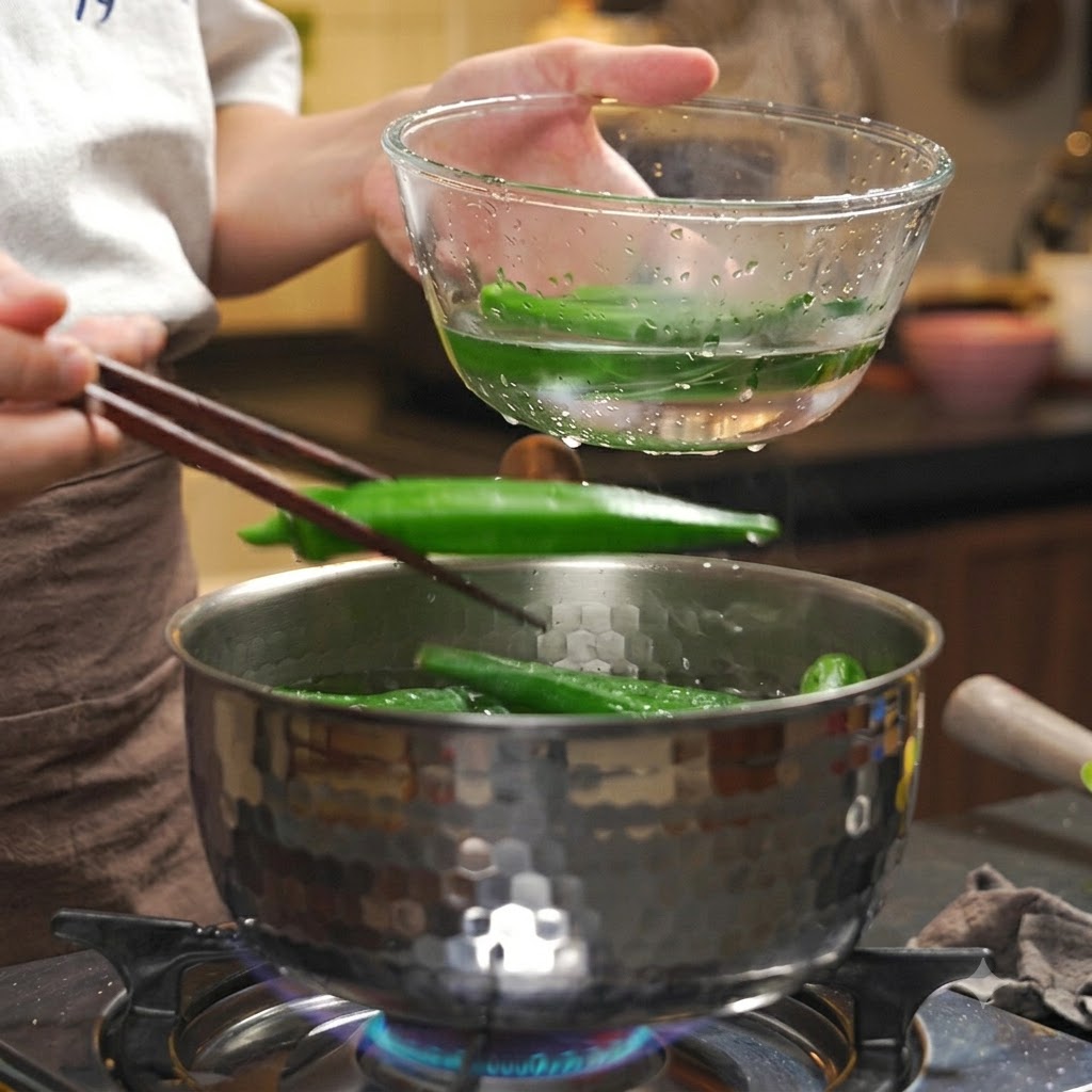 Cooked okra being transferred with chopsticks from a hot pot into a clear bowl of ice water.