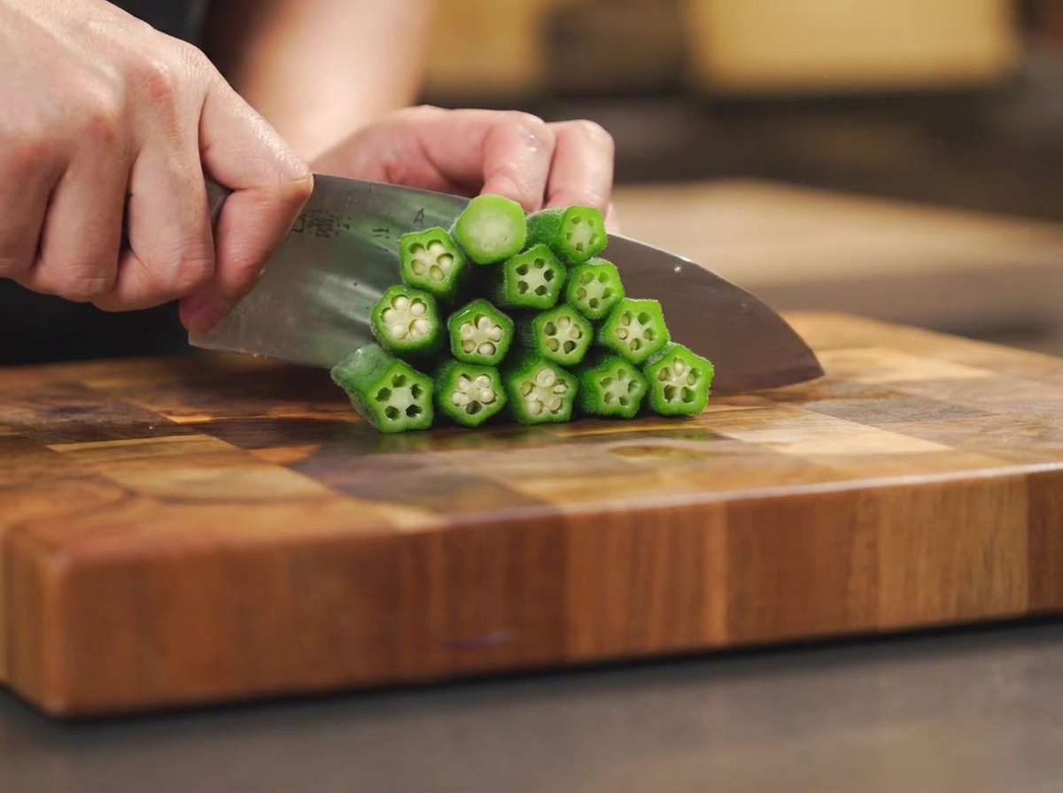 A chefs knife cutting the stems and halving cooked okra pods stacked on a wooden cutting board.