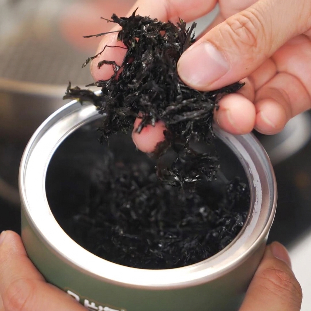 Hands holding dark shredded dry seaweed above a heated stainless steel pan.