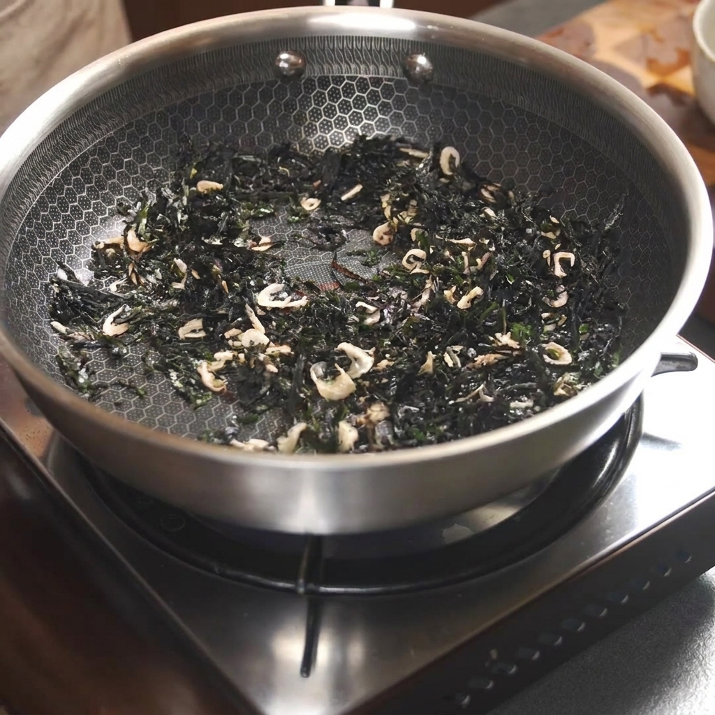Dried seaweed and small dried shrimp being stir-fried inside a pan using chopsticks.