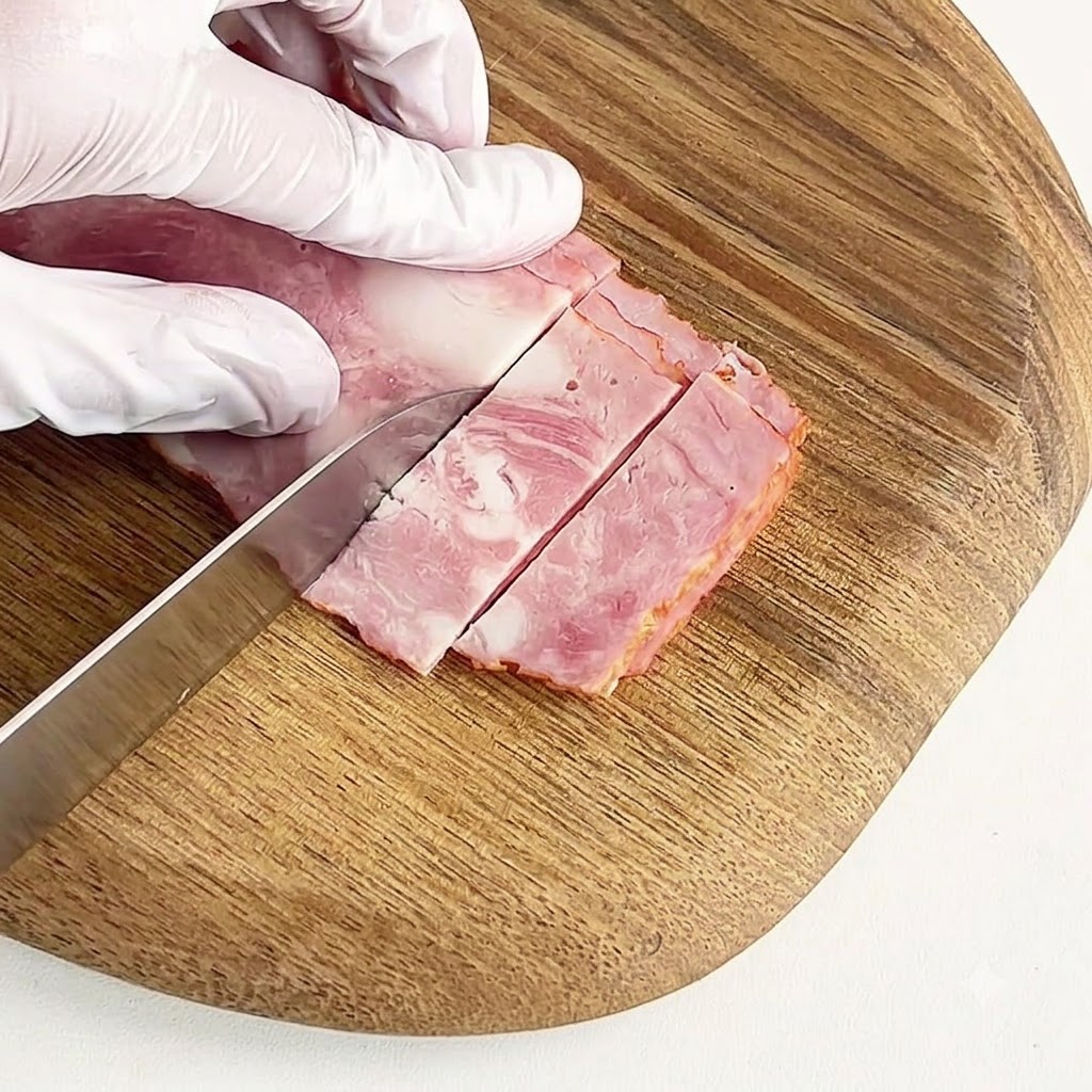 A gloved hand slicing cured bacon into small pieces on a round wooden cutting board.