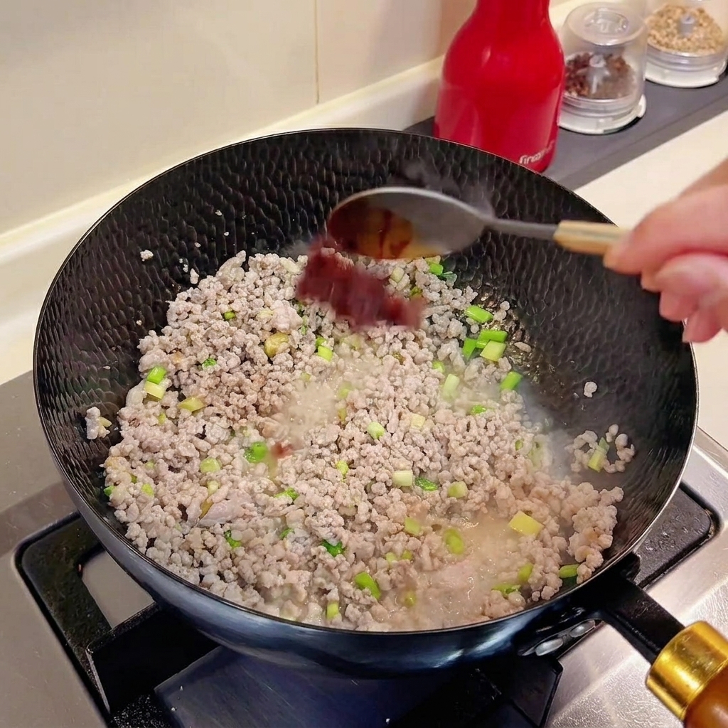 A spoonful of doubanjiang being added into a wok containing minced meat and aromatics.
