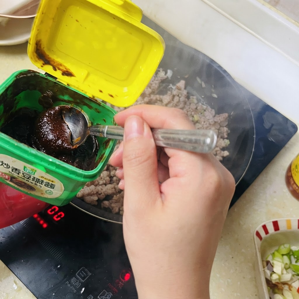 A spoonful of dark bean paste being scooped from a green container into the stir-fried minced pork.