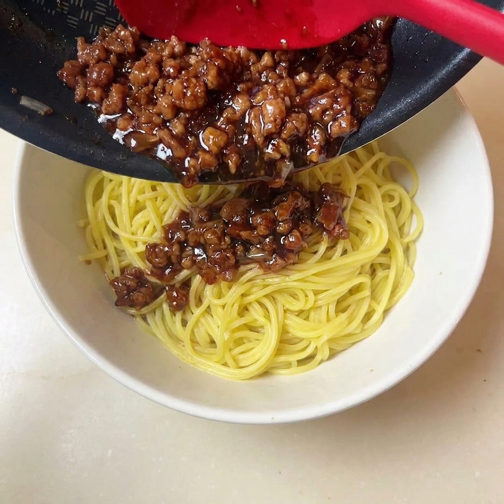 A red spatula spooning rich, dark minced meat sauce onto a bowl of yellow noodles.