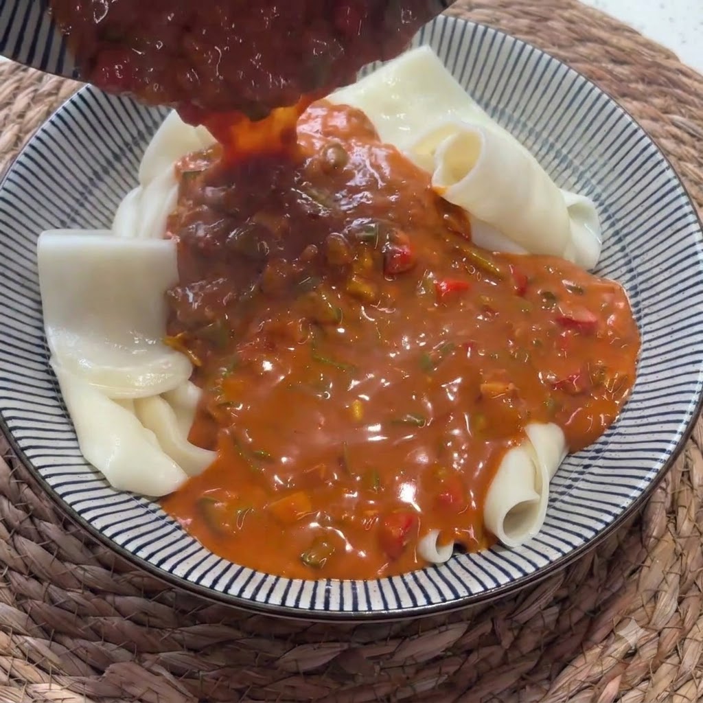 Rich red sauce being poured from a ladle onto a bowl filled with folded wide noodles.