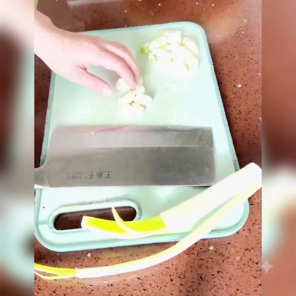 Scallion, ginger, and garlic being chopped and minced on a green cutting board with a large cleaver.