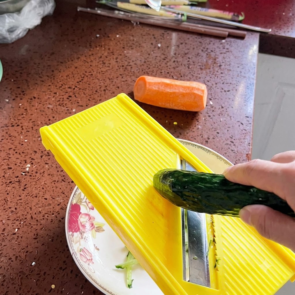 A fresh cucumber being grated into thin shreds using a bright yellow mandoline slicer over a decorative plate.