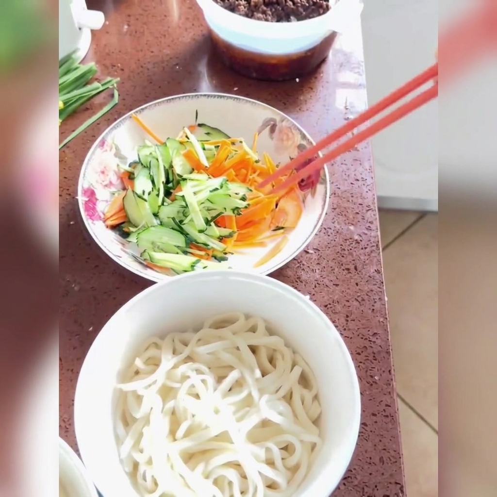 Fried sauce being spooned and drizzled over freshly cooked noodles topped with shredded cucumber and carrots in a white bowl.