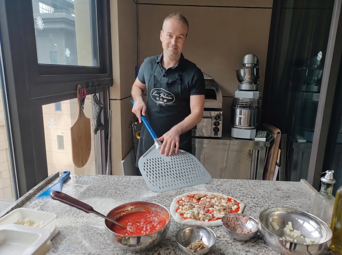 A pizza maker holding a large perforated metal pizza peel positioned right beside a fully assembled raw pizza.