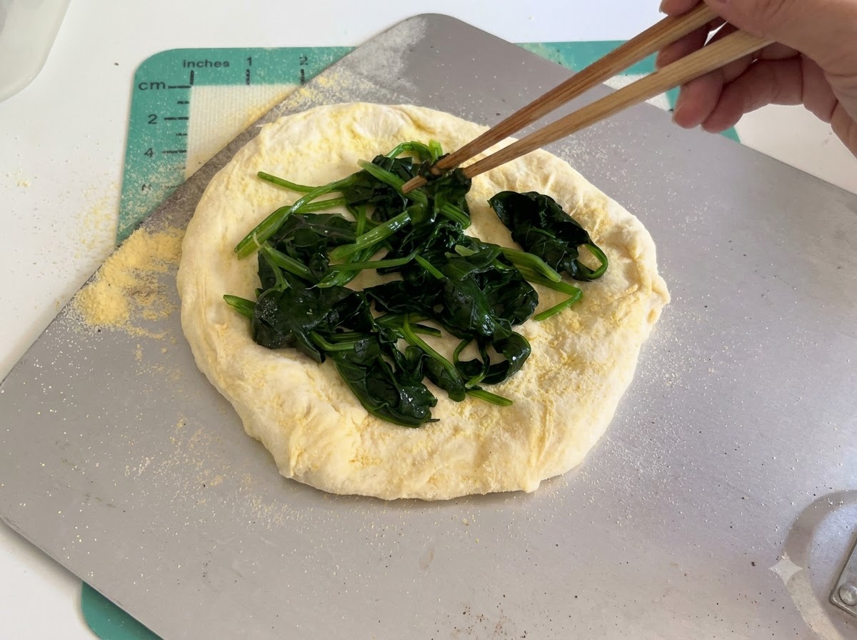 White cheese slices being carefully arranged over the spinach layer on top of raw pizza dough using chopsticks.