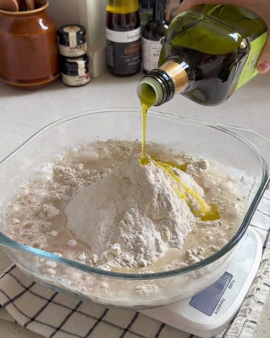 Olive oil being poured over a mound of flour and water mixture inside a clear glass bowl on a kitchen scale.