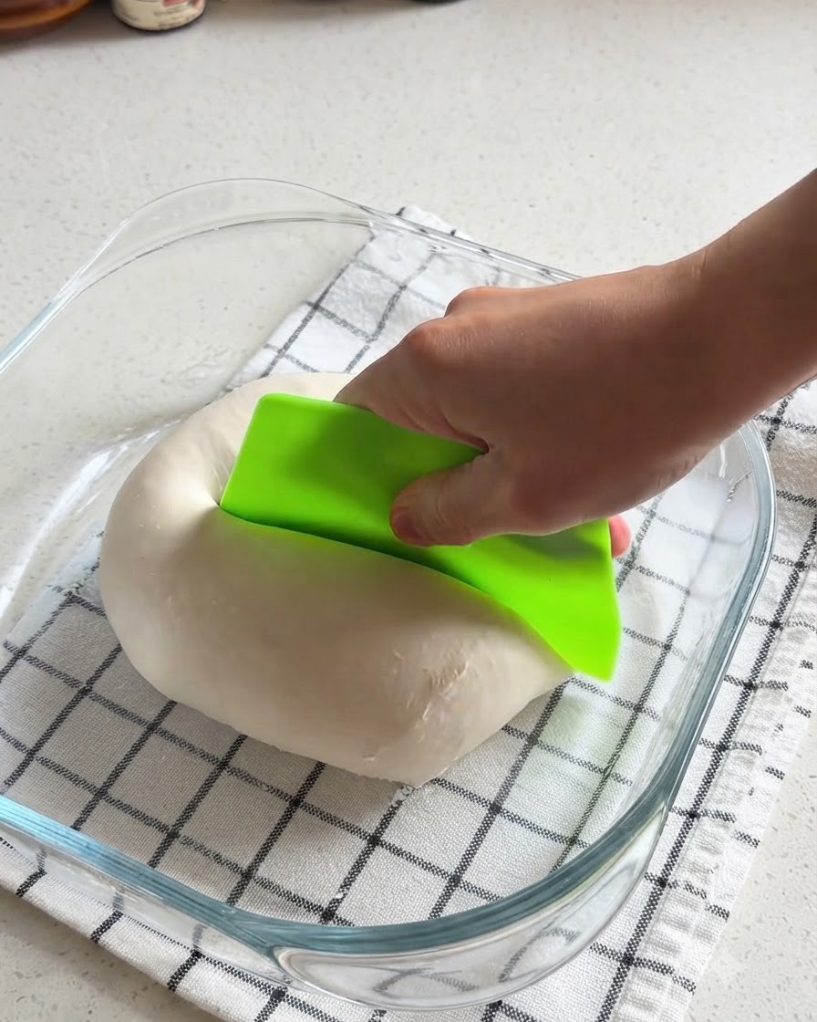 A hand using a bright green bench scraper to cut a smooth ball of pizza dough in half inside a glass dish.