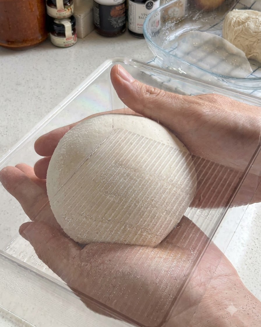 Two hands holding a perfectly rounded ball of pizza dough as it is placed into a clear plastic fermentation container.