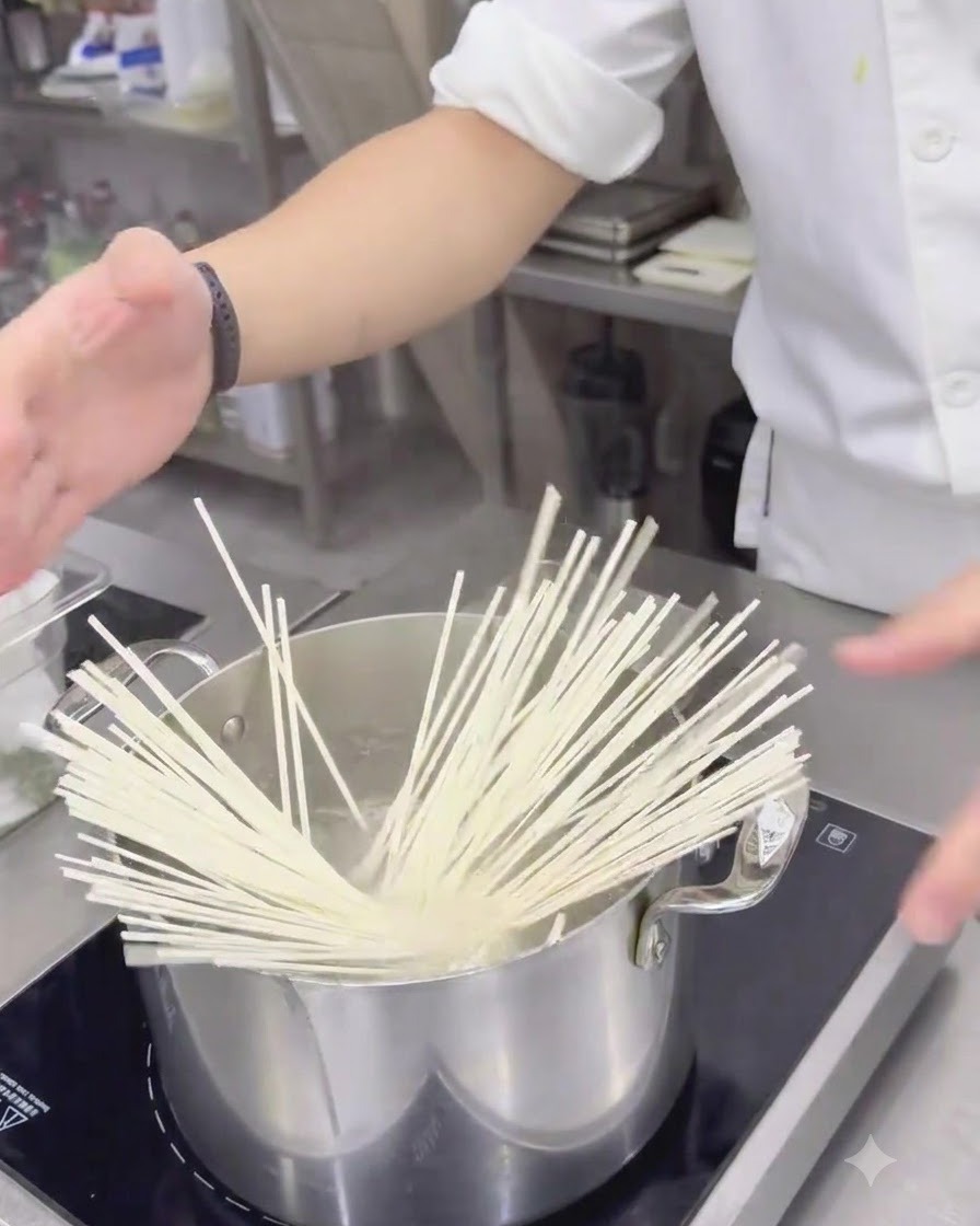 Dried white udon noodles being dropped into a stainless steel pot of boiling water.