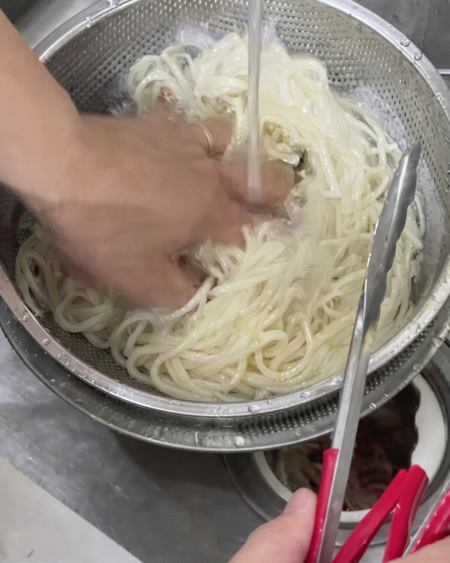 A hand tossing cooked udon noodles in a metal strainer under cold running tap water.