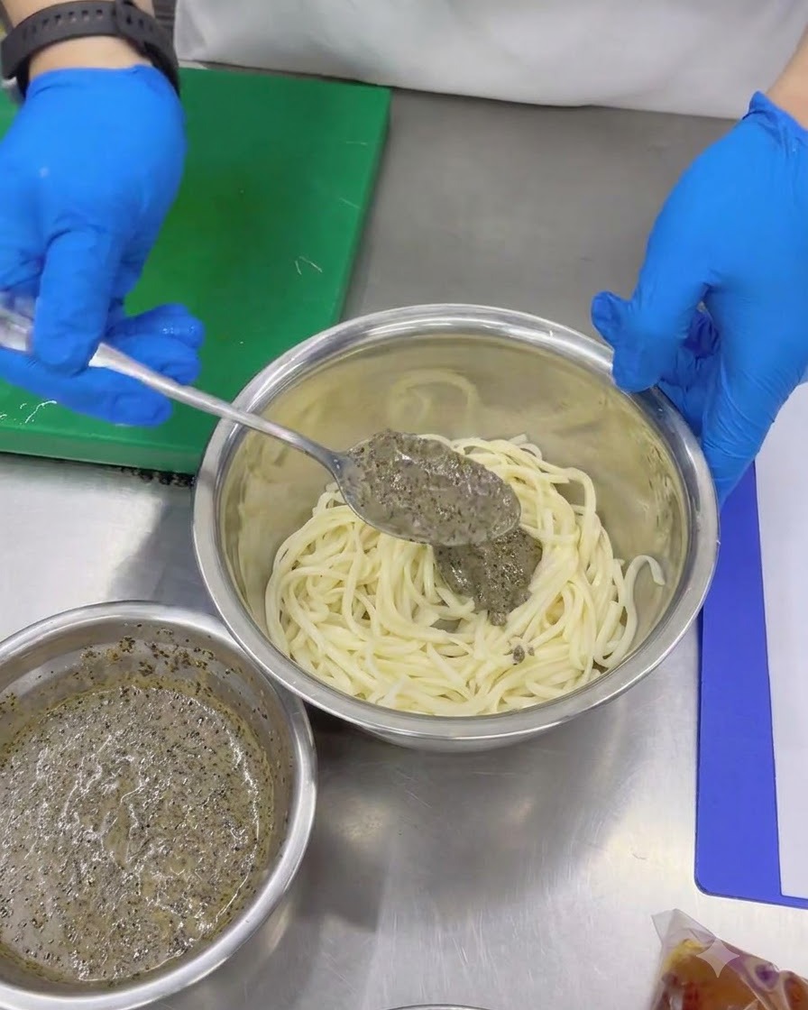 Gloved hands using a spoon to add thick black truffle sauce to cold udon noodles in a metal mixing bowl.