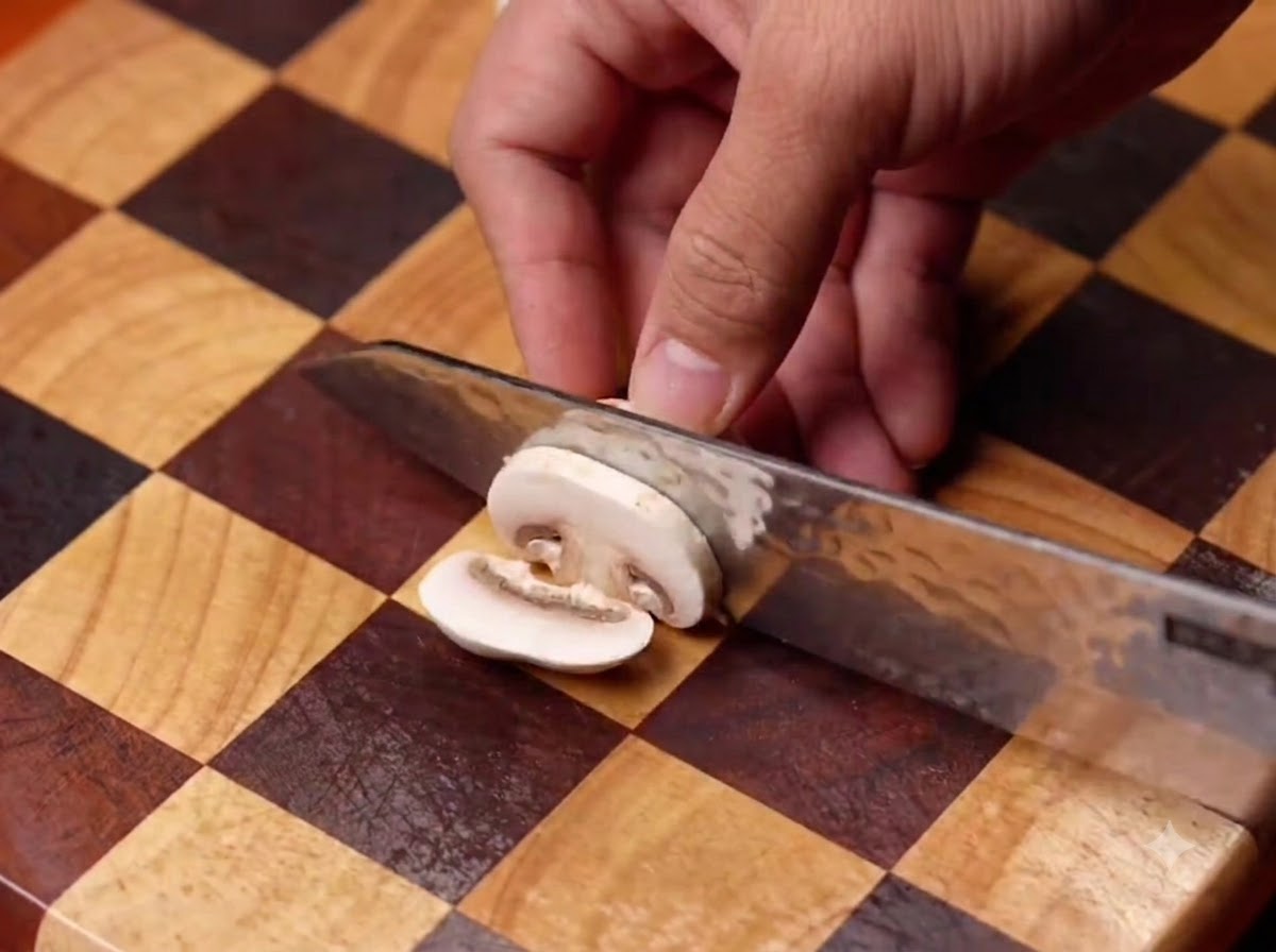 Button mushrooms being sliced with a knife on a checkered wooden cutting board, then added into a black pan.