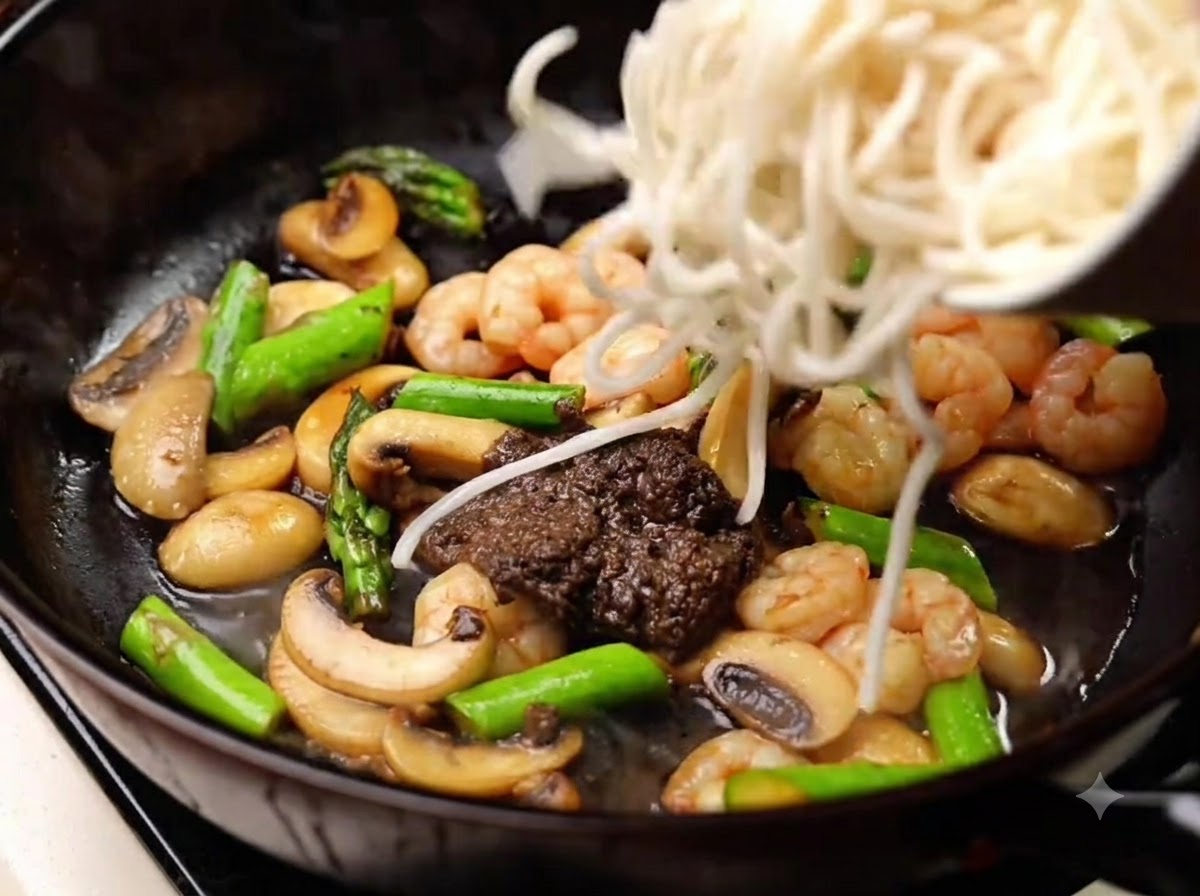 Cooked noodles being added into the pan to be mixed with the sauce, mushrooms, shrimp, and asparagus.