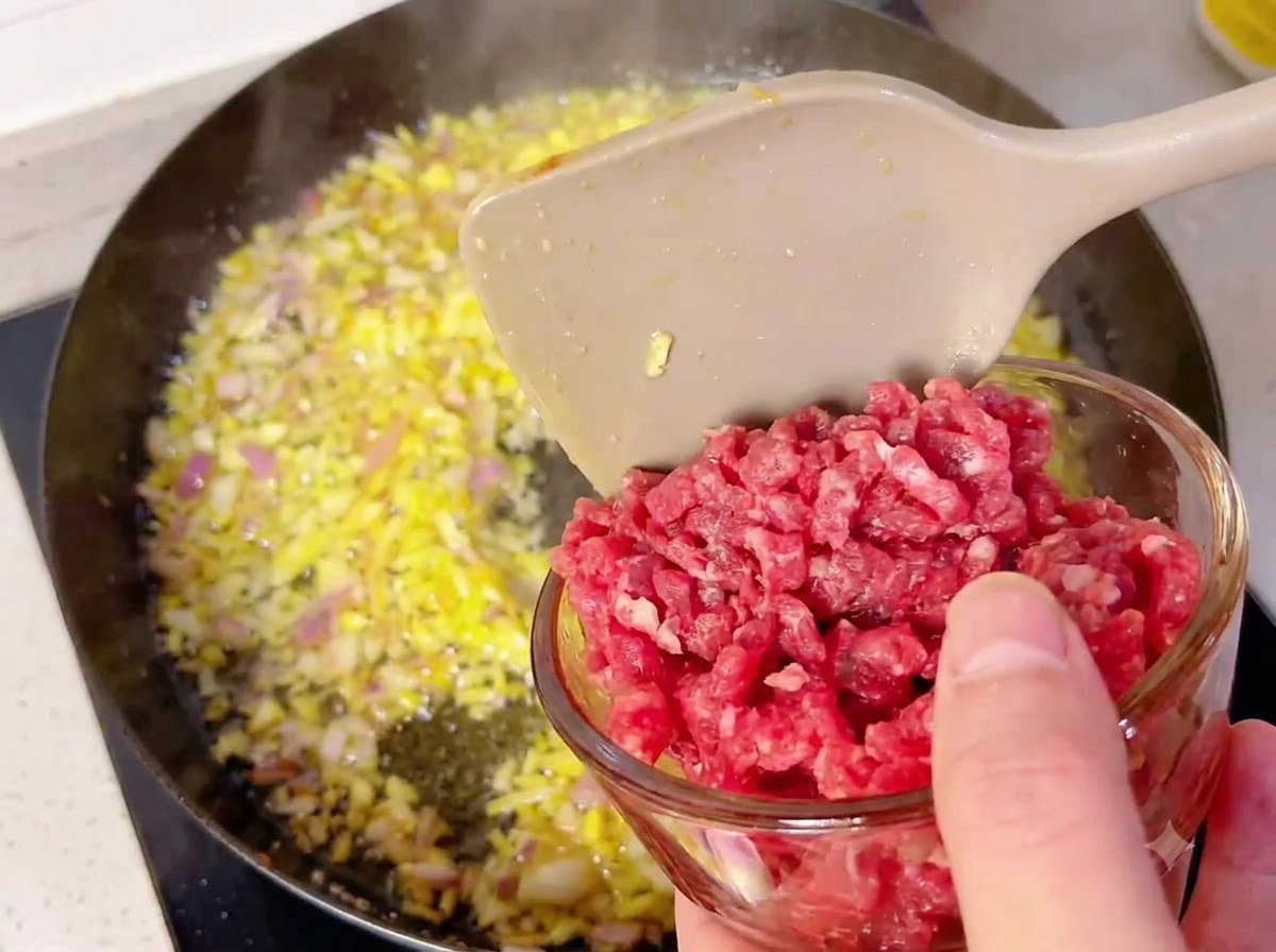A small glass bowl pouring beef cubes into a hot pan filled with sautéed onions and garlic.