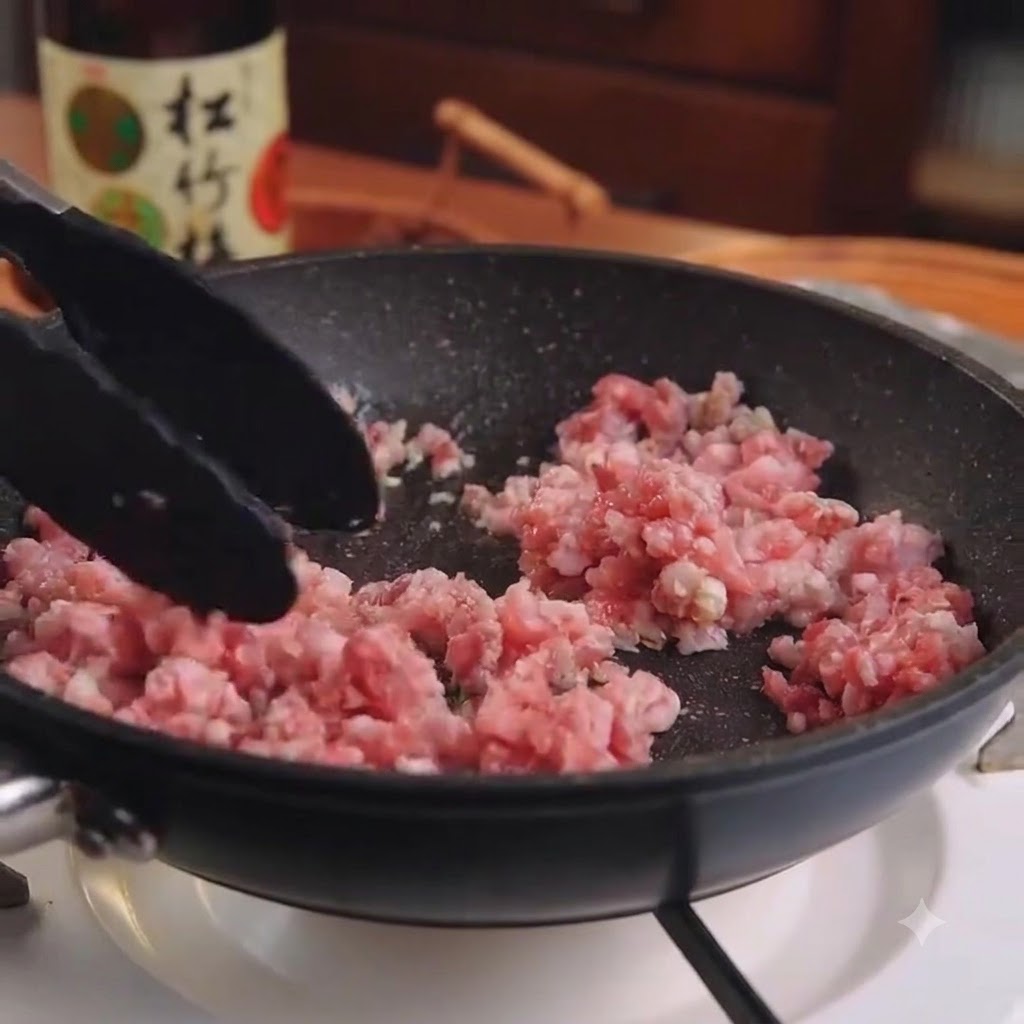 Raw minced pork and garlic being stirred in a black frying pan with tongs.