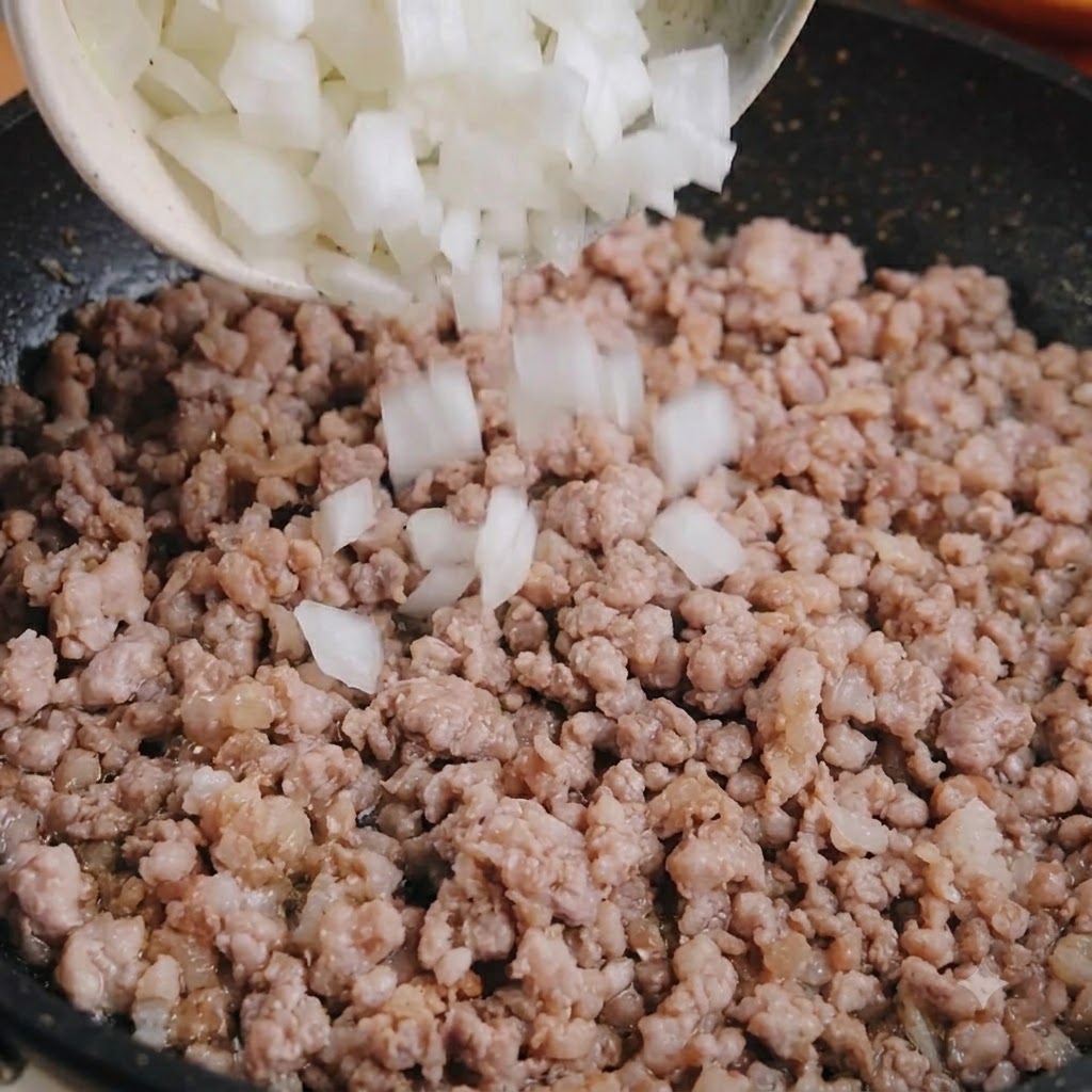 Diced white onion being poured from a bowl into a pan of cooked minced pork.