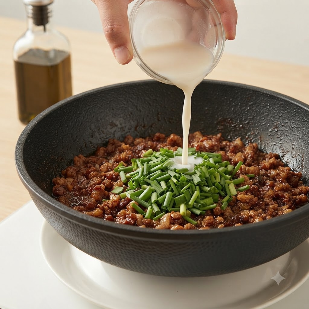 A hand pouring a white starch slurry from a small glass bowl into a pan of minced meat and chives.