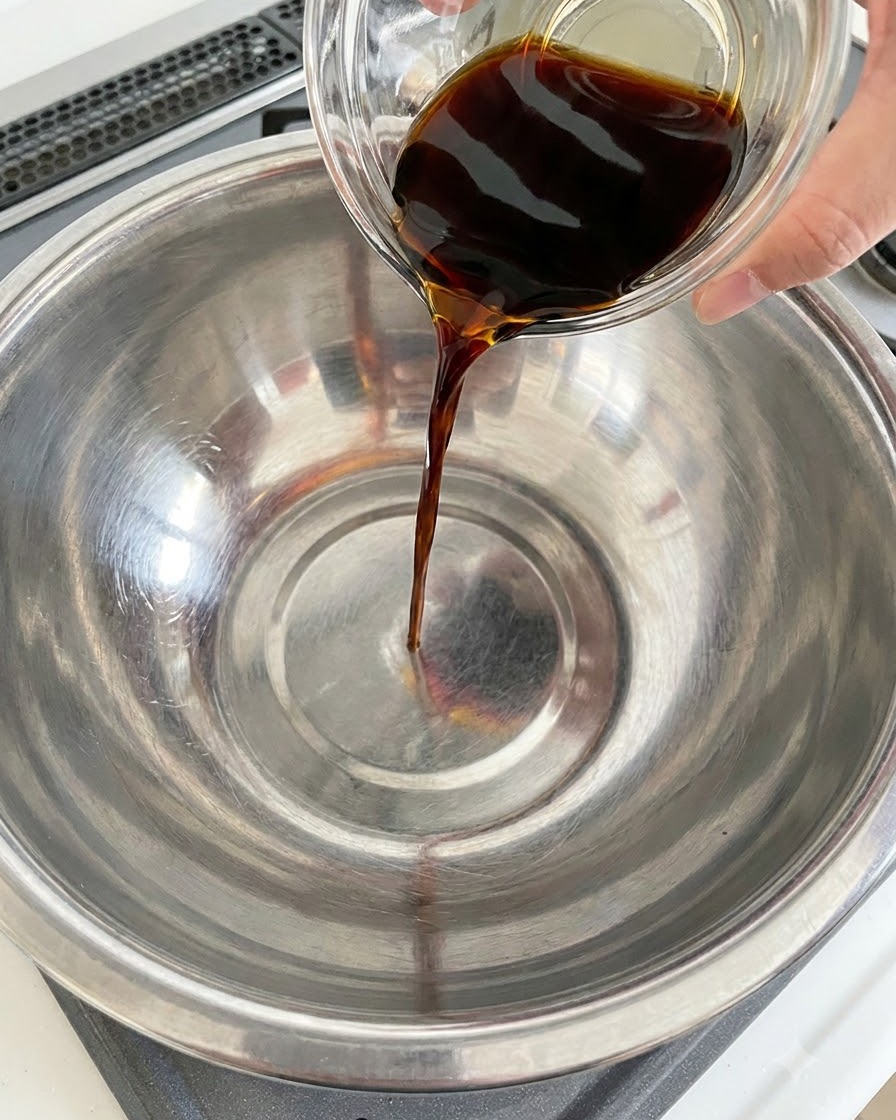 Dark soy sauce being poured from a glass container into a stainless steel bowl.