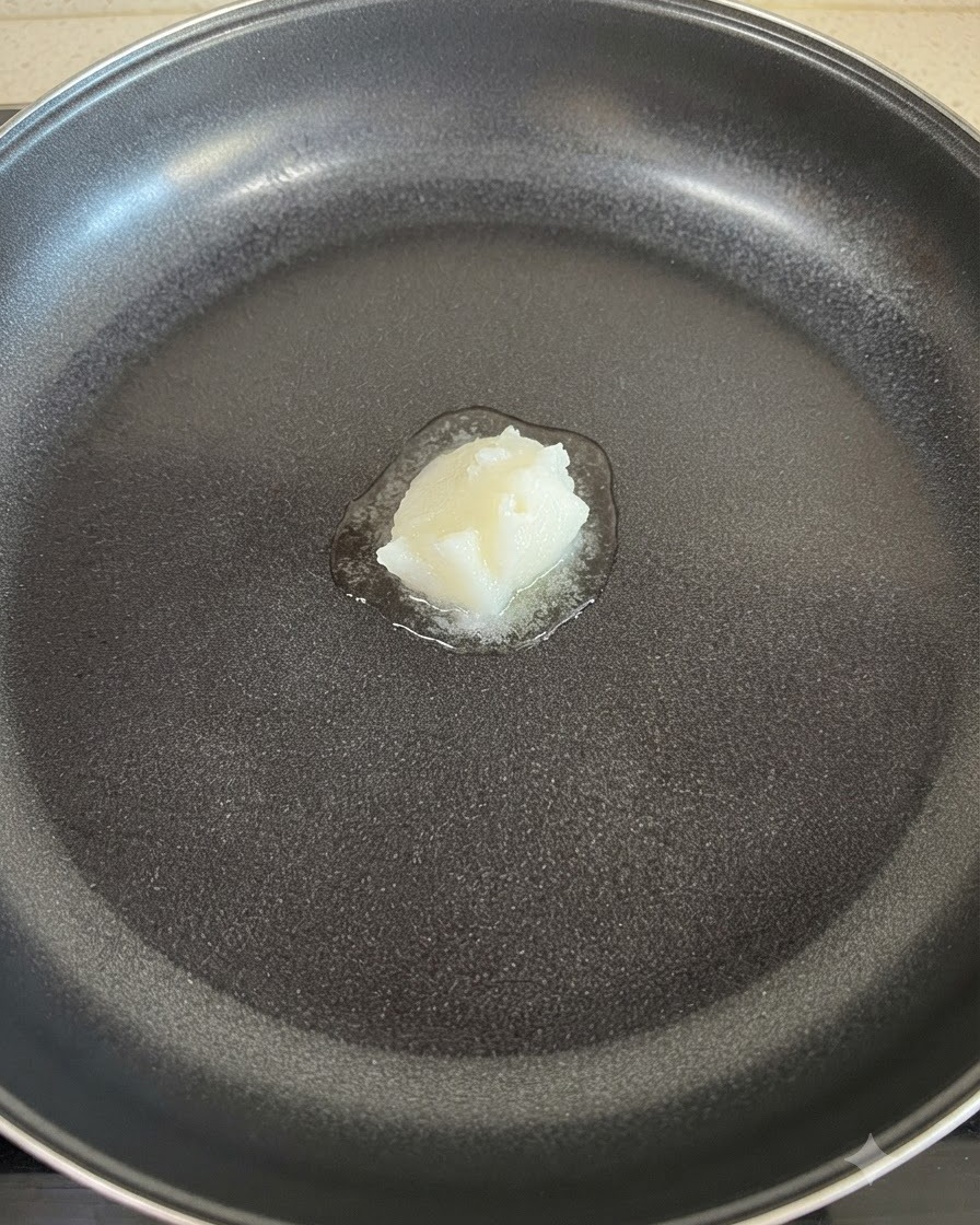 A solid dollop of white lard melting in the center of a non-stick frying pan.