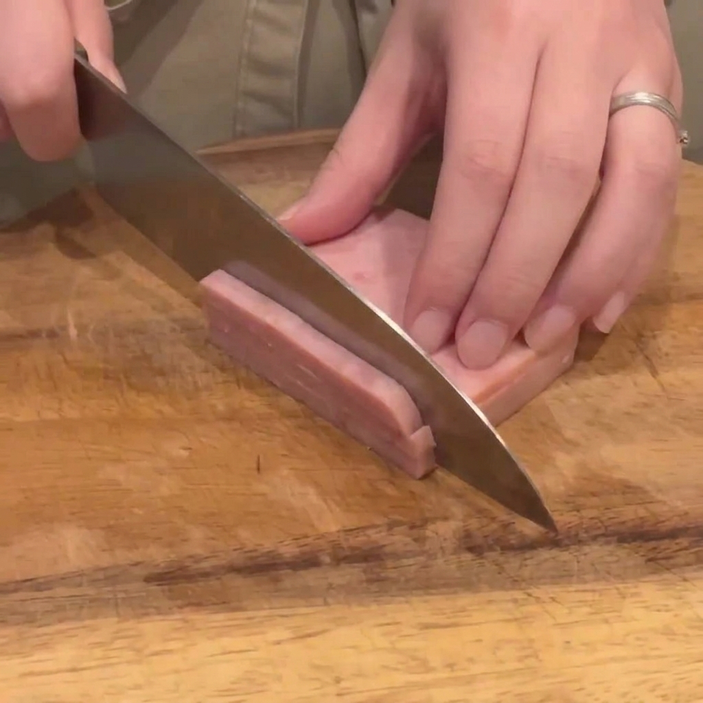 A kitchen knife slicing a block of pink ham into thin, even strips on a wooden board.