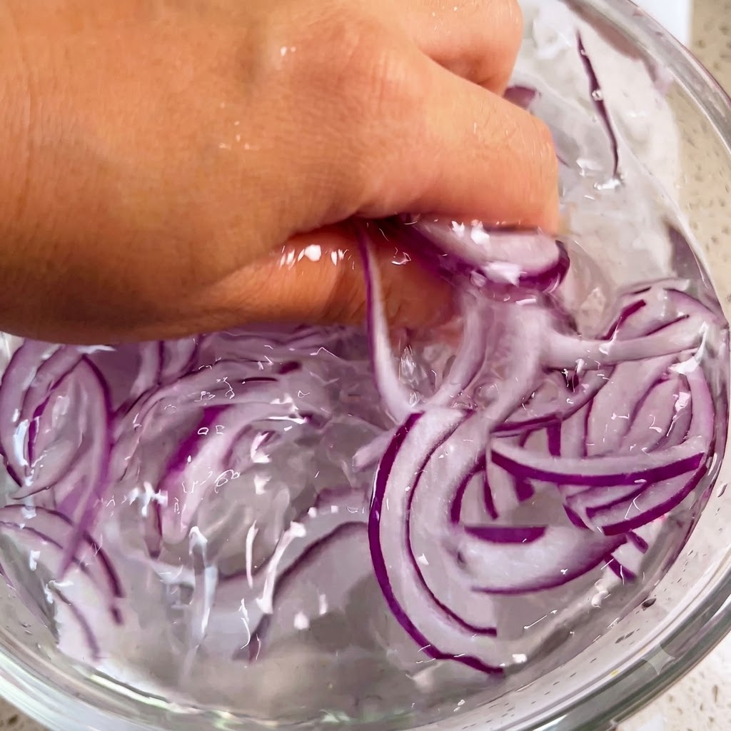 A hand gently washing purple onion shreds inside a bowl filled with ice water.