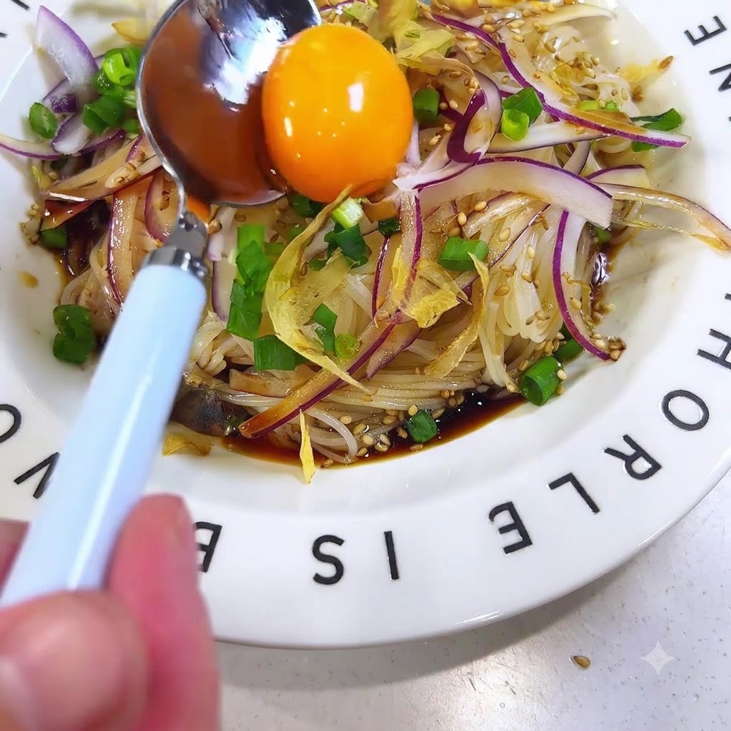 A bright raw egg yolk resting on a spoon being positioned in the center of the dressed somen noodles.