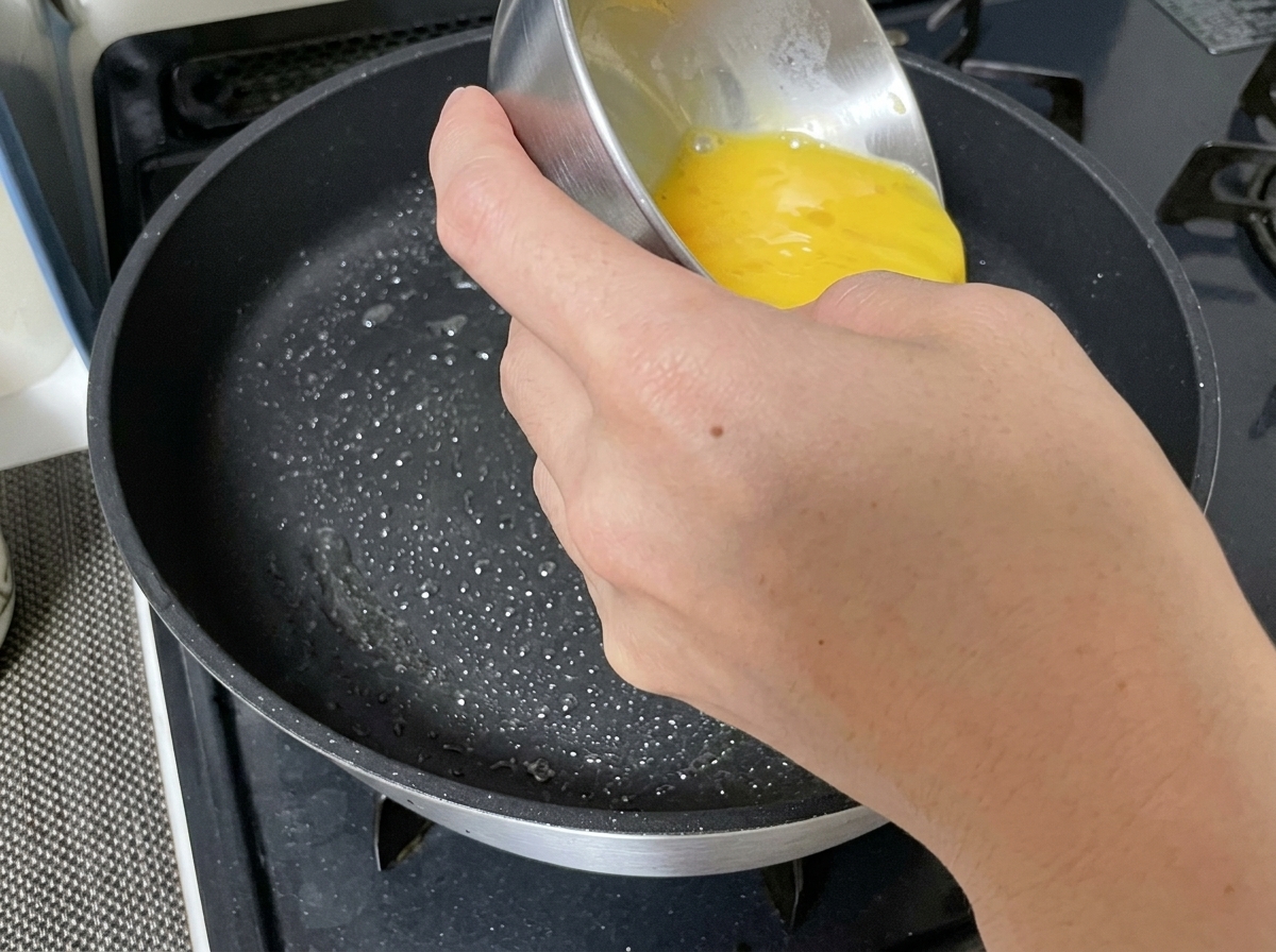 Beaten egg liquid being poured into a greased frying pan.