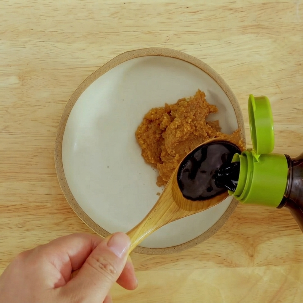 Dark soy sauce being poured into a bowl with thick red miso paste.