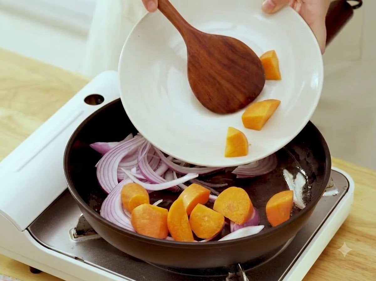 Raw carrot chunks being added from a plate into a pan containing sautéed onions.