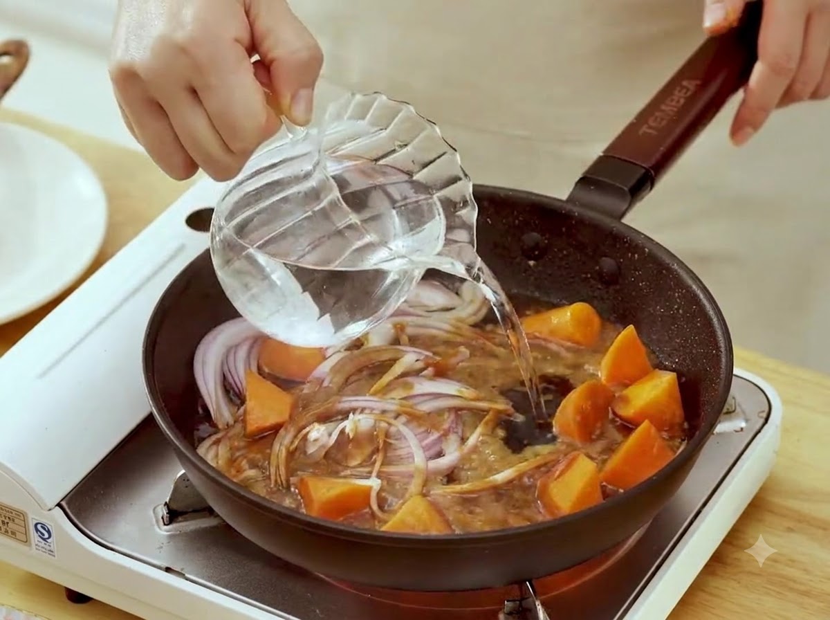 Water being poured from a glass pitcher into a frying pan with sliced red onions and carrot chunks in miso sauce.