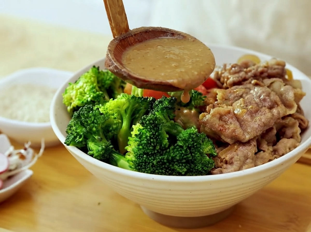 A wooden ladle pouring thick brown miso sauce over a bowl filled with broccoli, cooked beef, and steamed rice.