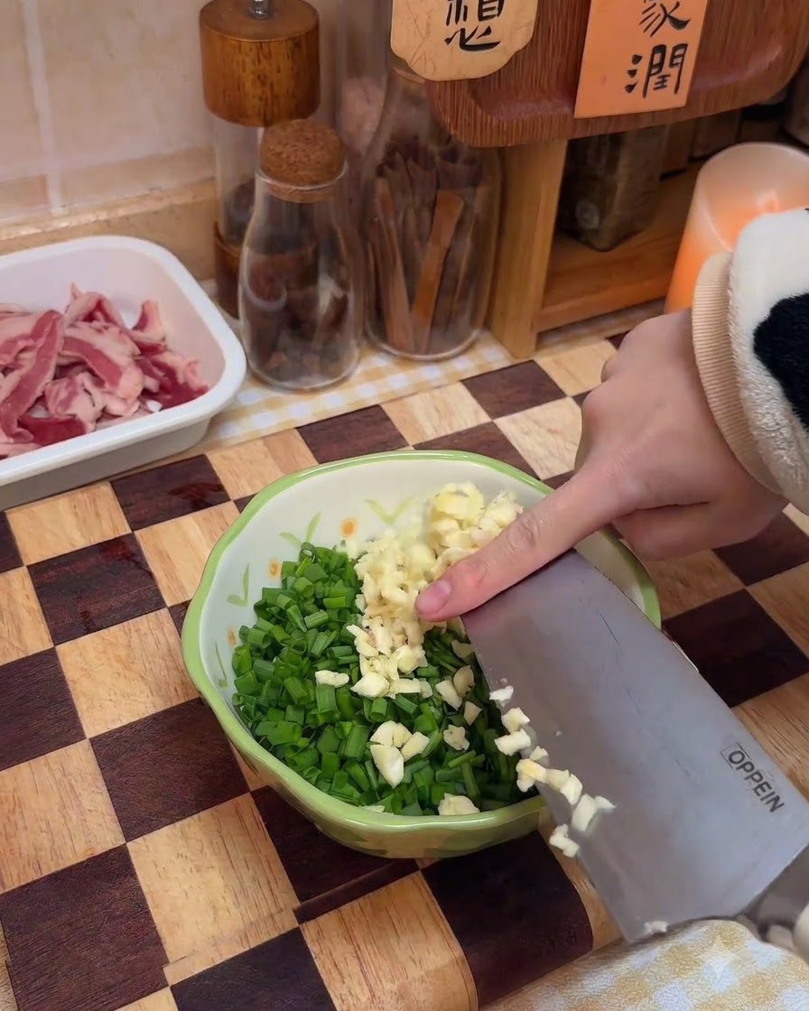 Minced garlic being added to a bowl containing chopped green onions and spices.