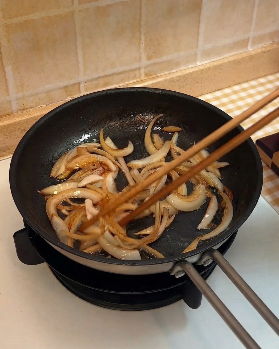 Onion strips being stir-fried in a pan using wooden chopsticks.