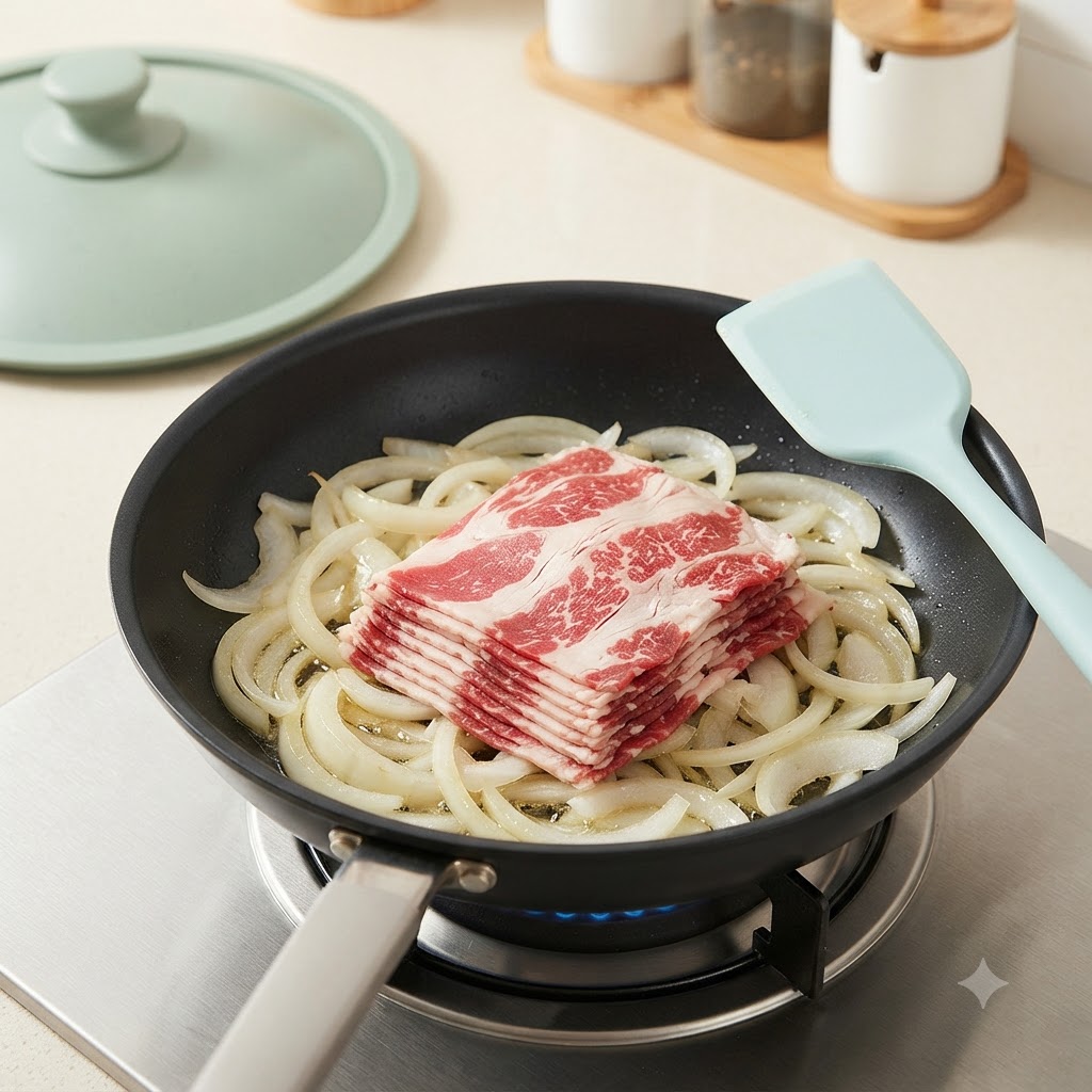 Sliced beef pieces laid over the sautéed onions in a frying pan on the stove.