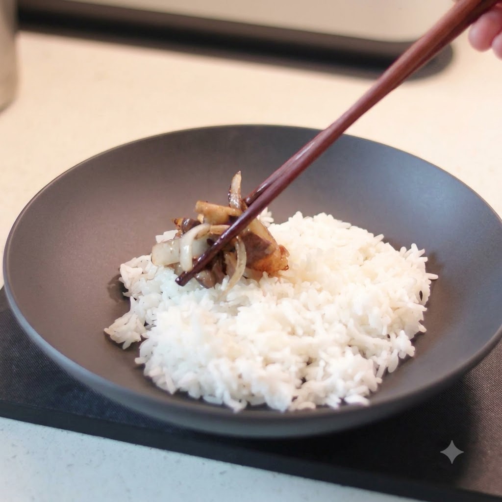 A pair of wooden chopsticks holding a portion of cooked beef and caramelized onions, placing it onto a dark ceramic bowl filled with white rice.
