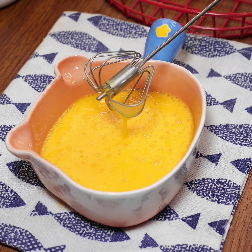 A blue-handled small whisk blending yellow eggs inside a pink bear-shaped bowl.