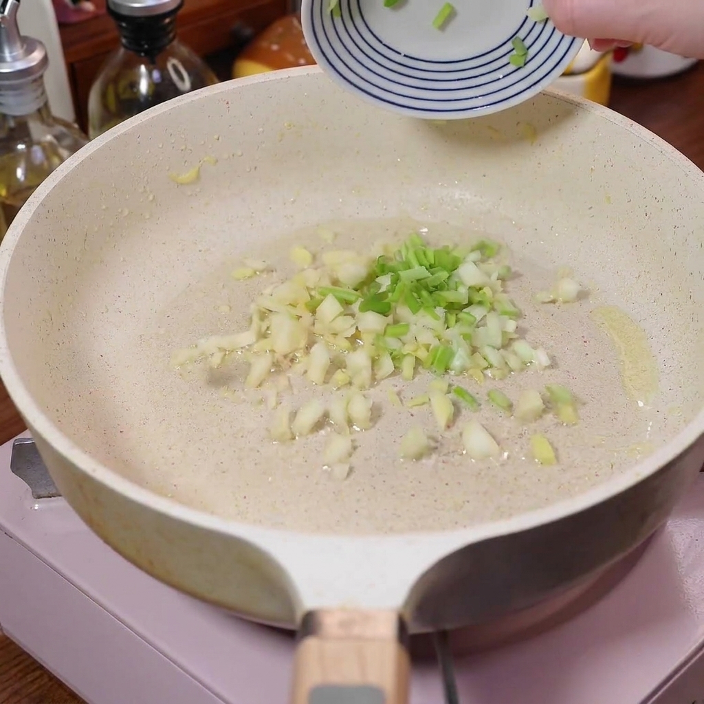 Minced green onions, garlic, and ginger sizzling in a white pan.