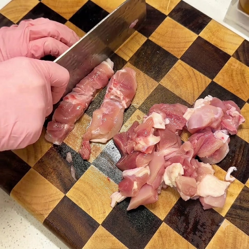 Hands wearing pink gloves using a cleaver to dice raw chicken thighs on a checkered wooden cutting board.