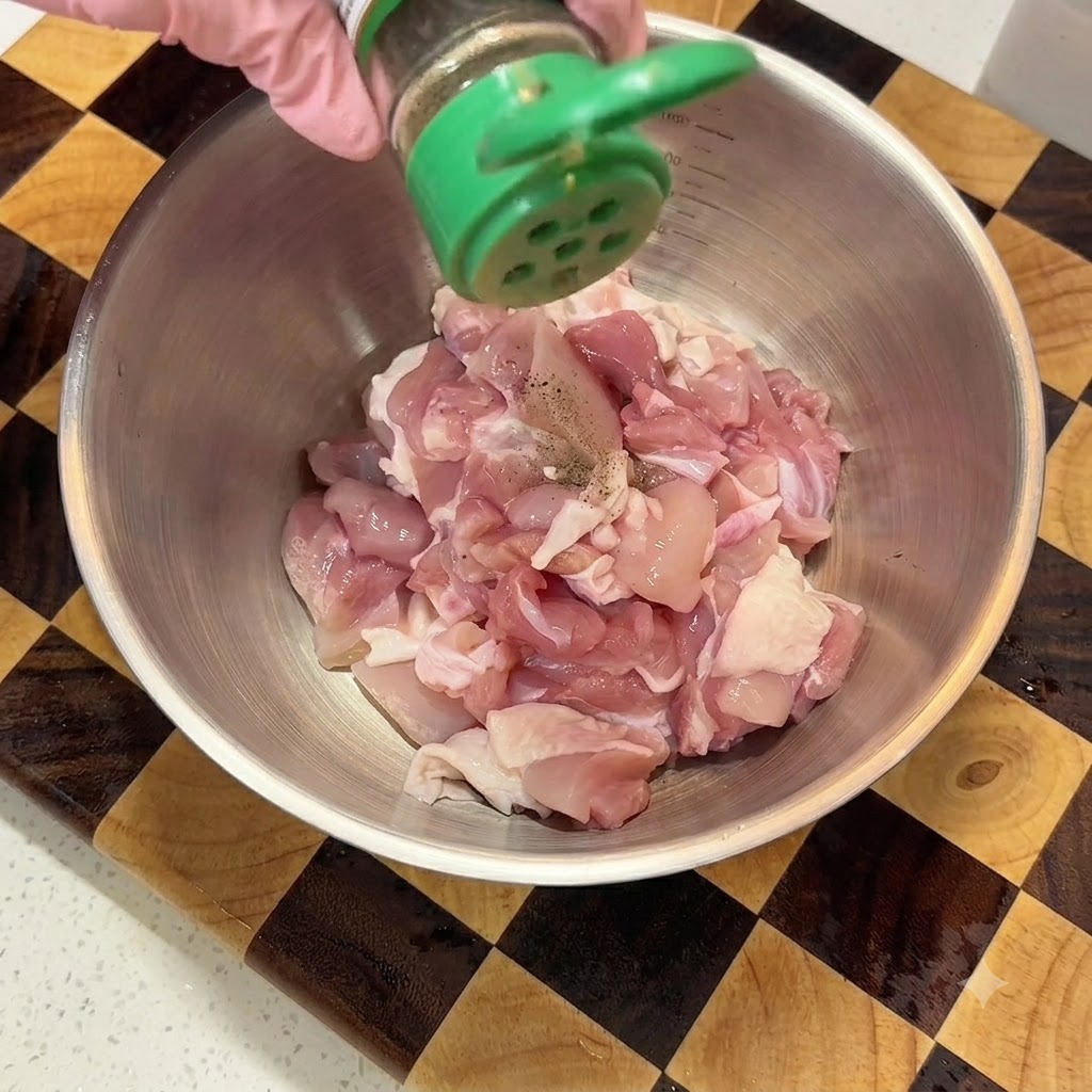 Sprinkling black pepper powder from a green shaker onto diced raw chicken in a metal mixing bowl.