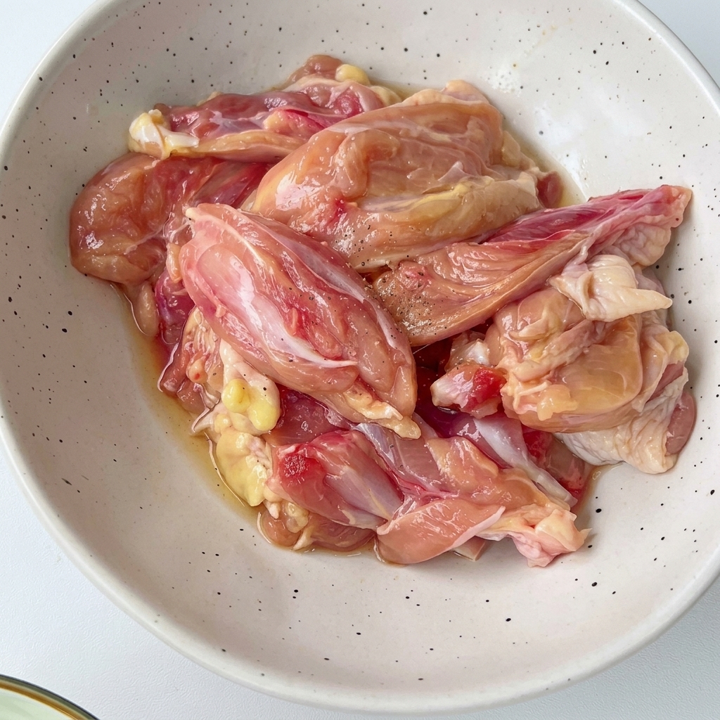 Chopped chicken pieces inside a white speckled bowl mixed with seasonings and aromatics.