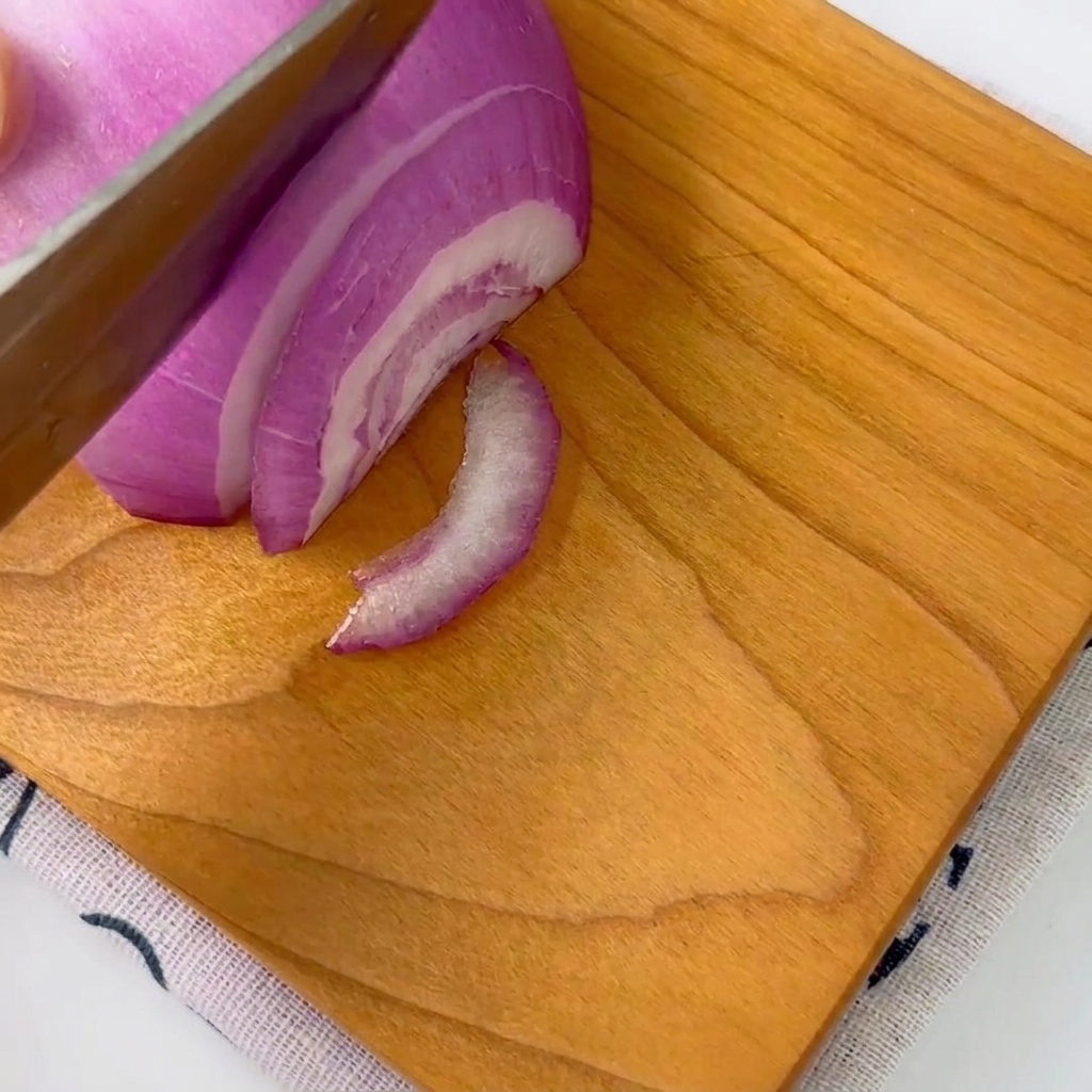 A red onion being carefully sliced into thin strips on a wooden cutting board with a knife.