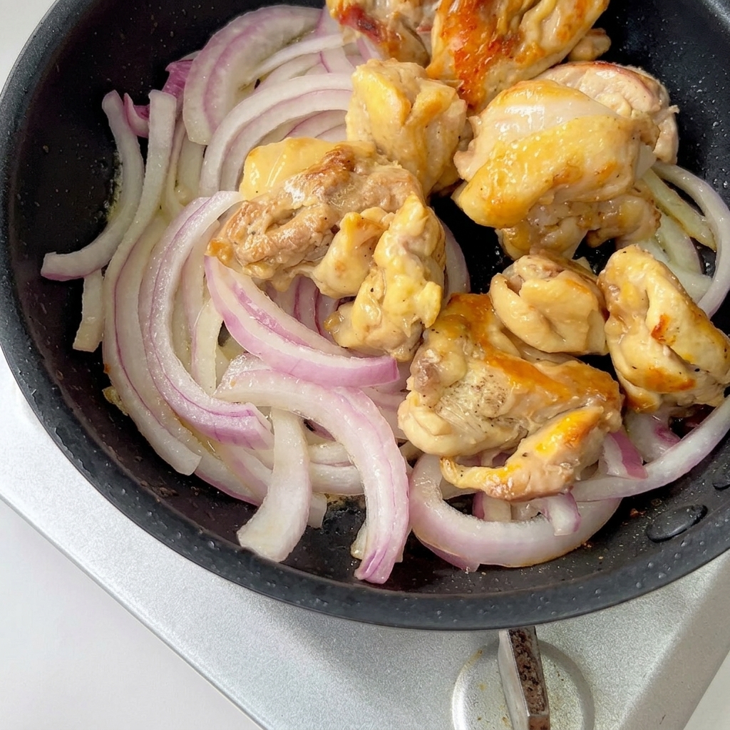 Cooked chicken pieces and sliced red onions simmering in a pan with sauce.