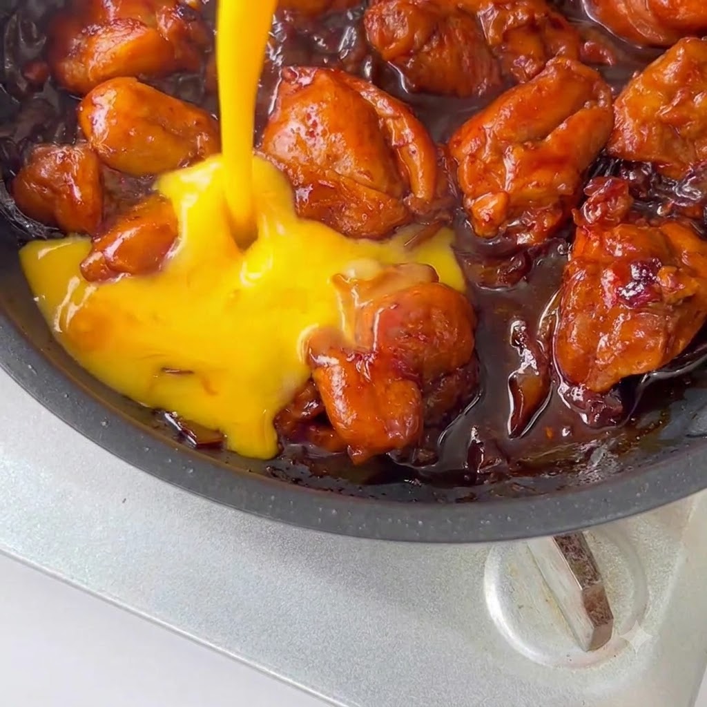 Beaten eggs being poured over chicken and sauce in a skillet.